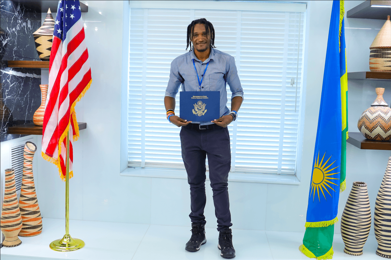 Thomas Shiva holding a fellowship certificate between the United States and Rwanda flags.